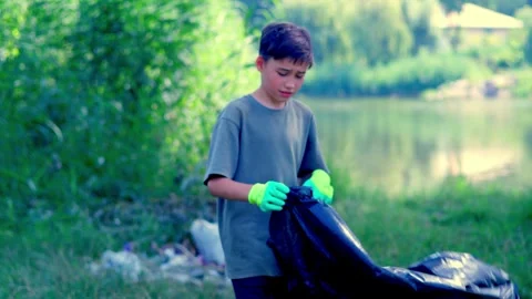 Boy carrying large black garbage bag across green riverbank Stock Footage 340464295