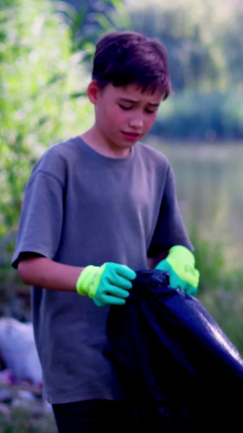 Boy carrying large black garbage bag across green riverbank Stock Footage 340577538