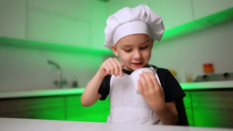 Boy in chef hat holding spoon and plastic pot of yogurt in green kitchen Stock Footage 342162486