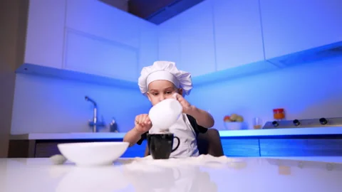 Boy in chef hat pouring flour into mug. A child cooks at a white table in a Stock Footage 343382321