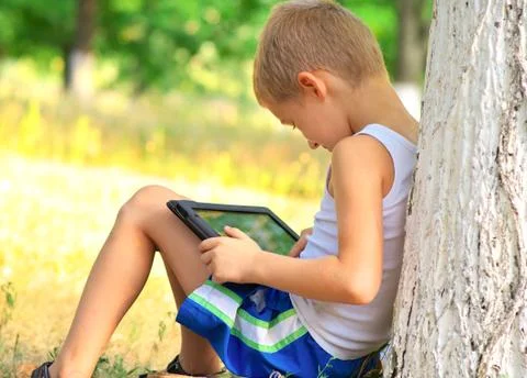 Boy child playing with tablet pc sitting on skateboard outdoor with forest on Stock Photos