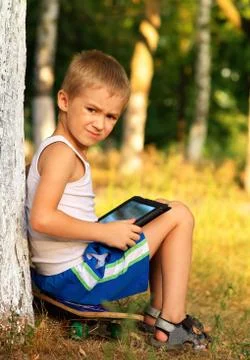 Boy child playing with tablet pc sitting on skateboard outdoor with forest on Stock Photos