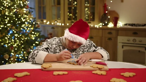 Boy coloring gingerbread man in blue color in kitchen with illuminated xmas tree Stock Footage 256796981