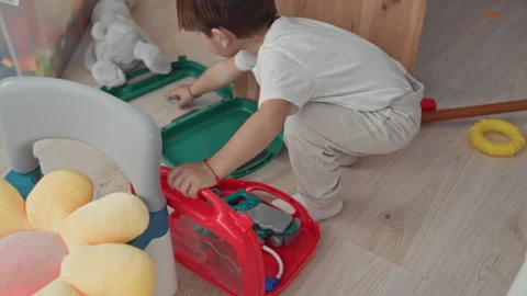 Boy crouches with hand reaching into red toy medical kit on playroom floor .. Stock Footage 339249165