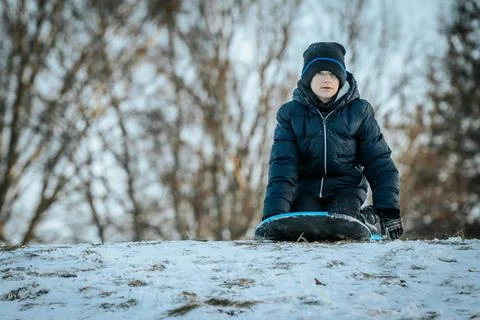 A boy in a dark winter jacket and hat sleds down a snowy hill on a blue sled Stock Photos