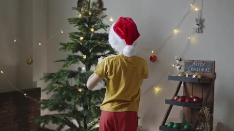Boy decorates a Christmas tree by hanging Christmas toy balls in the room Stock Footage 291605149