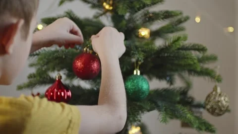 Boy decorates a Christmas tree by hanging Christmas toy balls in the room Video stock 291605735