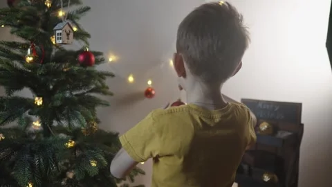 Boy decorates a Christmas tree by hanging Christmas toy balls in the room Stock Footage 291607442