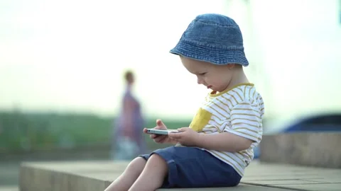 Boy in denim hat and striped t-shirt sits outdoors using smartphone.  Stock-Footage 337006299
