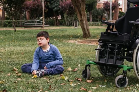 Boy with a disability sitting on a grass in a park with a wheelchair near him. Stock Photos