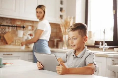 The boy does his homework in the kitchen using a tablet online, talking with the Stock Photos