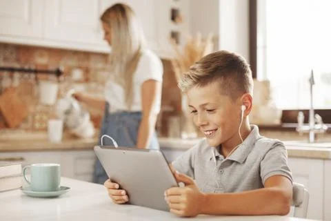 The boy does his homework in the kitchen on the tablet online, talking with the Stock Photos