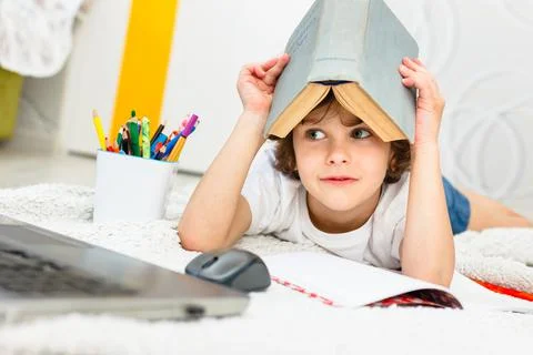Boy does homework and holds a book on his head in light room. Stock Photos