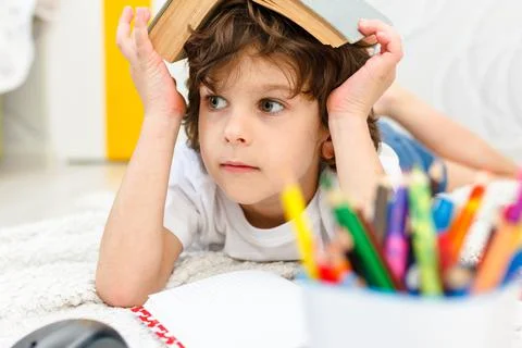 Boy does homework and holds a book on his head in light room. Stock Photos