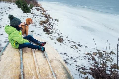 Boy with dog sitting together near frozen lake Stock Photos