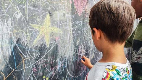 A boy draws with colored chalks on a board with drawings. A large board with  Stock Photos