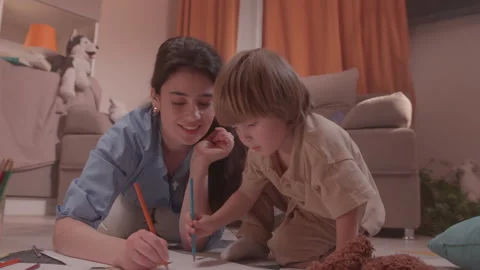 Boy draws with orange pencil while woman watches on floor Stock Footage 324912128