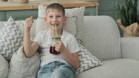 Boy drinking fresh juice sitting at sofa with pillows. Happy boy in white shirt Vídeo Stock 107065660