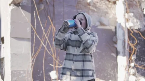 A boy drinks tea from a thermos against the backdrop of a ruined house, Stock Footage 232663767