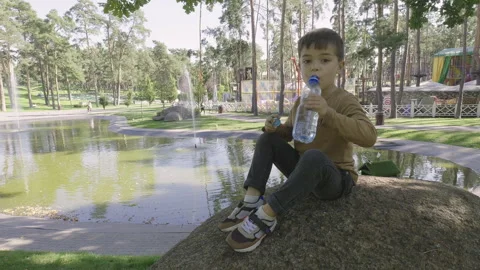 A boy drinks water from a bottle while sitting on a stone in the park. Stock Footage 251831416