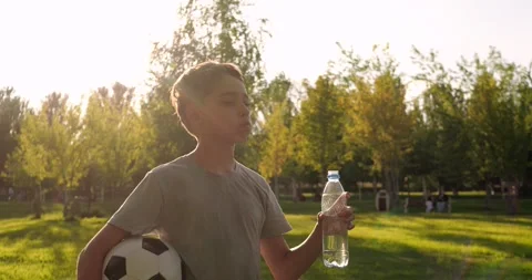 Boy drinks water holding soccer ball in sunny park. Child refreshes after Stock Footage 346690616