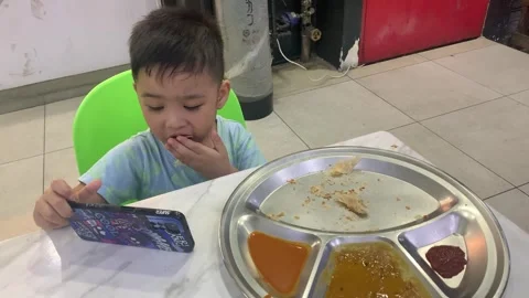 A boy eating roti canai while looking at his mobile phone Stock Footage 285836782