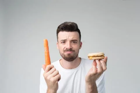Boy eats an carrot and hamburger. Man makes the choice between fast and healthy 库存照片
