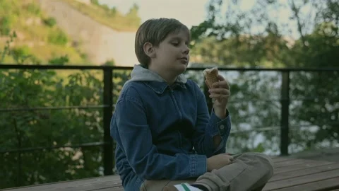 Boy eats ice cream cone sits on wooden bench by river at sunset 库存影片 342194584