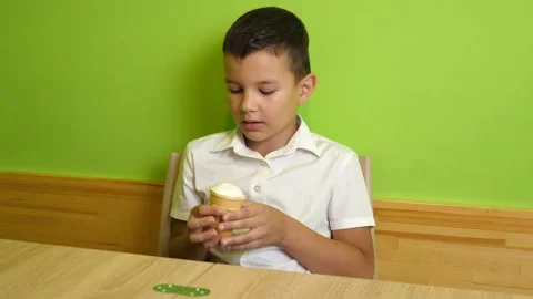 A boy eats ice cream in a waffle cone while sitting at the table and enjoying Stock Footage 338079093