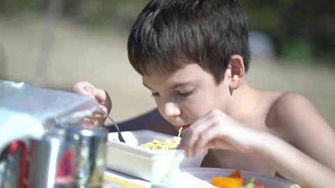 Boy eats instant noodles with a fork from a styrofoam box at an outdoor table Stock Footage 341989454