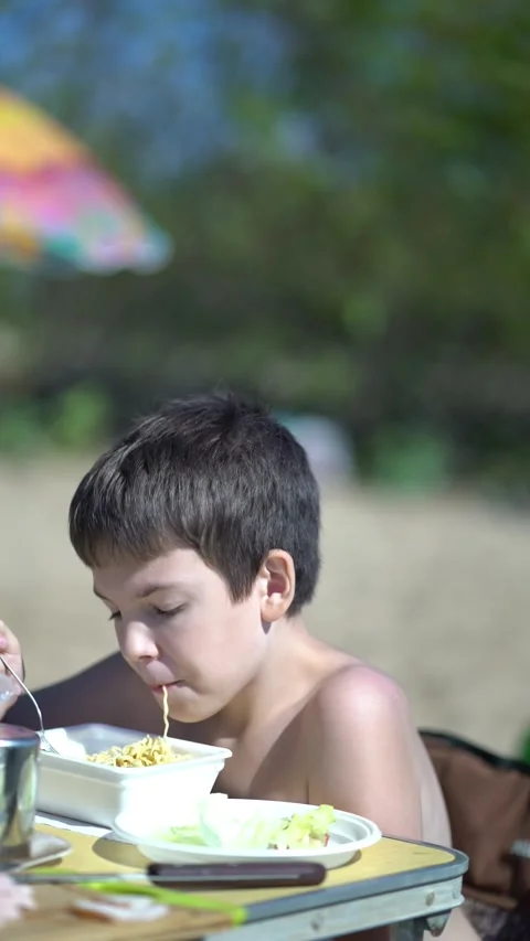 Boy eats instant noodles with a fork from a styrofoam box at an outdoor table Stock Footage 341989472