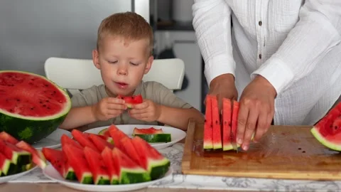 Boy eats a juicy slice of watermelon, mom's hands cut the watermelon Stock Footage 290982292