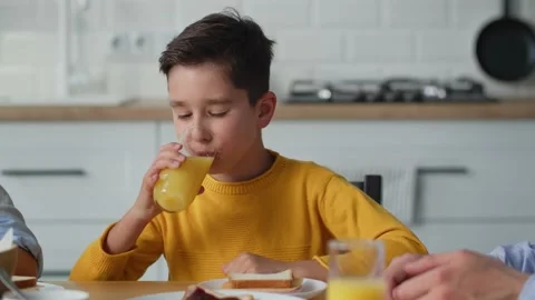 The boy eats a sandwich and drinks orange juice while sitting at the kitchen Stock Footage 240059507