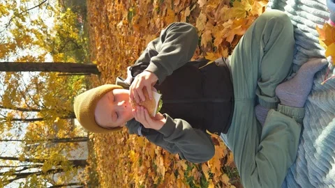 A boy eats a sandwich in an autumn park. Vertical footage. Stock Footage 288388736