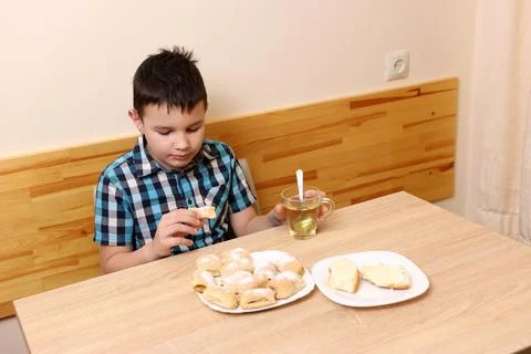 A boy eats sweet buns and drinks tea while sitting at the table. Stock Photos