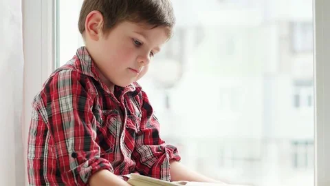 Boy flipping pages, little child wearing shirt sitting on window with old book Stock Footage
