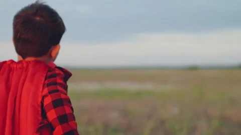 Boy with flowing red superhero raincoat runs along grassy meadow copying bird Stock Footage 258701197