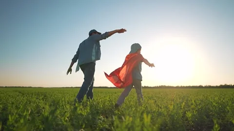 Boy in fluttering cape leads man across bright sunny field. Man stretches arms Stock Footage 346171627