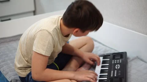 Boy fooling around playing black electronic piano synthesizer sitting on bed in  Video stock 307490471