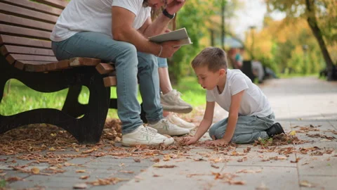 Boy gathering fall leaves next to adults on park bench during autumn showing Stock Footage 346723705