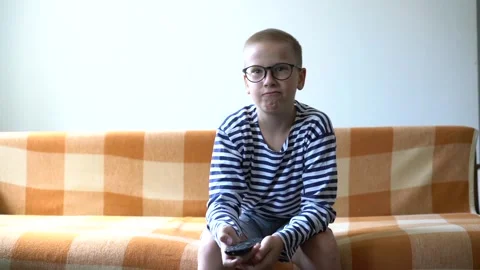 Boy with glasses holding remote control sitting on sofa watching tv Stock Footage 345212757