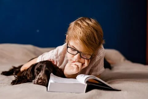Boy with glasses lying in bed reading with his dog by his side Stock Photos