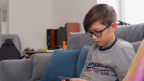 A boy with glasses sits on the couch and plays on the phone. Technology addicted Stock Footage 156608652