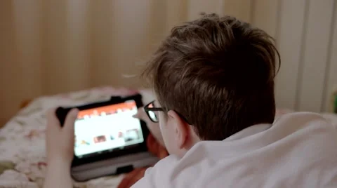 Boy in Glasses Watching Videos on a Tablet Lying on the Bed Vídeo Stock 62584576