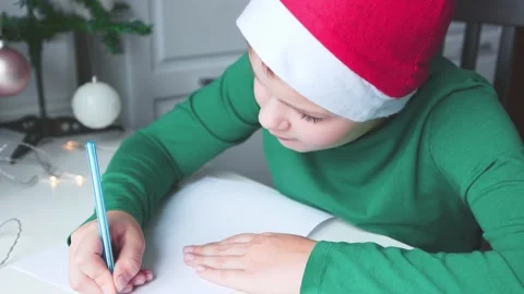 A boy in green clothes and a red cap sitting at the table writes Stock Footage 165384025