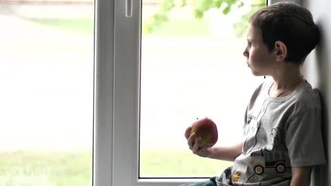 A boy happily eats a crisp, red apple and watching window Stock Footage 308643323
