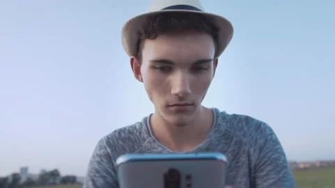 Boy in a hat reading messages on his cell phone, blue sky background, camera Stock Footage 170725796