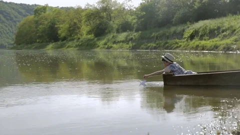 Boy in a hat, sitting in a boat, setting it on a paper boat swimming Stockbeeldmateriaal 76166481