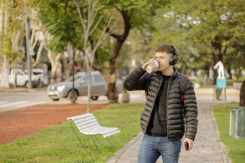 Boy with headphones drinks coffee while walking through a public park. 스톡 사진