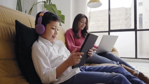 Boy with headphones playing with a tablet on the sofa at home while his mothe Stock Footage 150664752